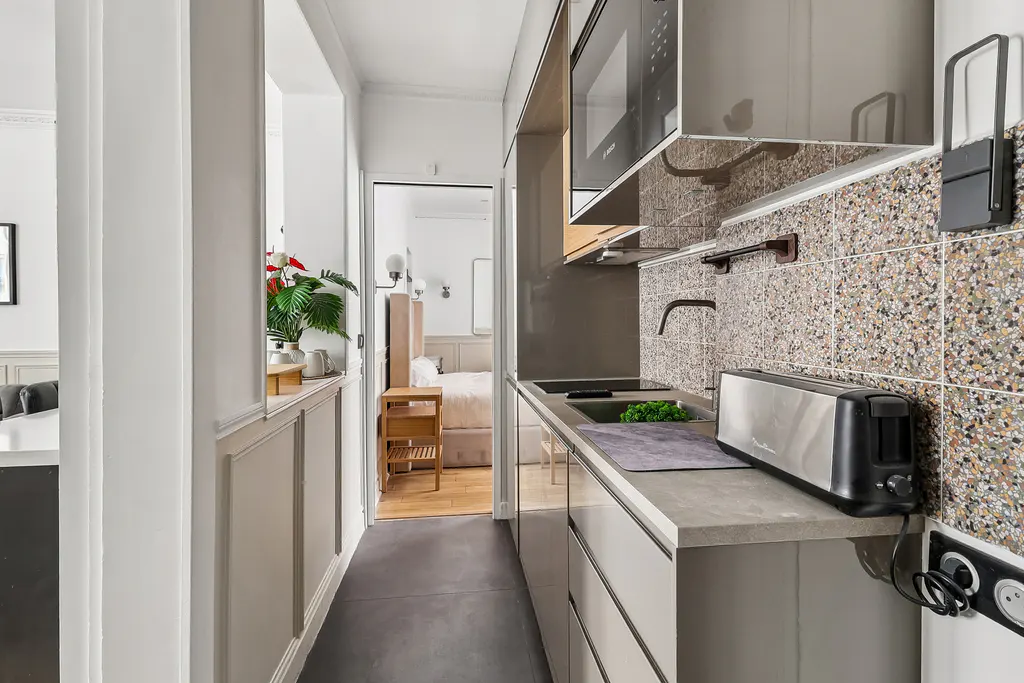 Modern galley kitchen with grey cabinets, terrazzo backsplash, and stainless steel appliances, leading to a bedroom.