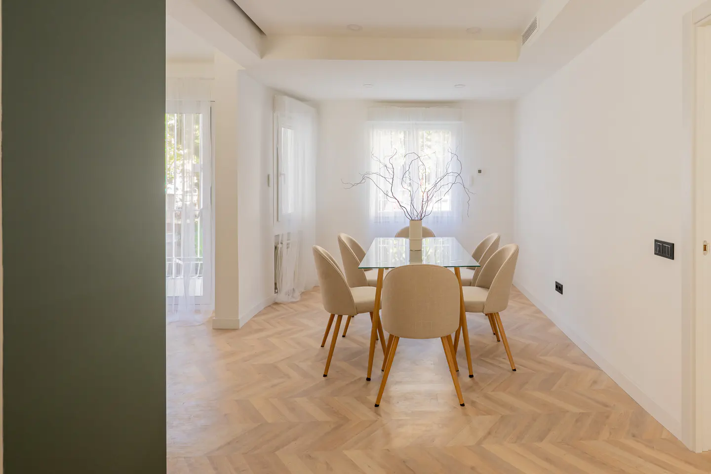 Bright dining room with a glass table, six beige chairs, and a vase with branches. Herringbone wood floors and white walls.