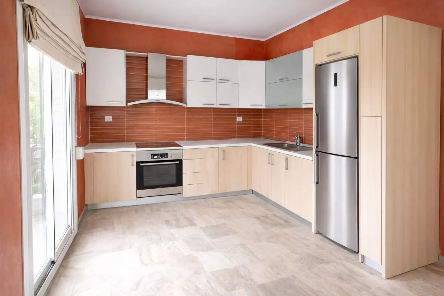 A modern kitchen with light wood cabinets, white upper cabinets, and stainless steel appliances. A large window with a beige shade is on the left. The walls are terracotta.