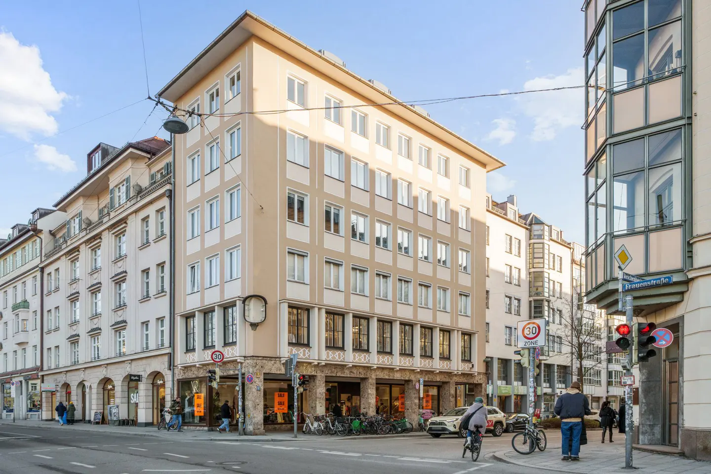 Street view of a beige building with many windows, shops on the ground floor, and people walking and cycling.