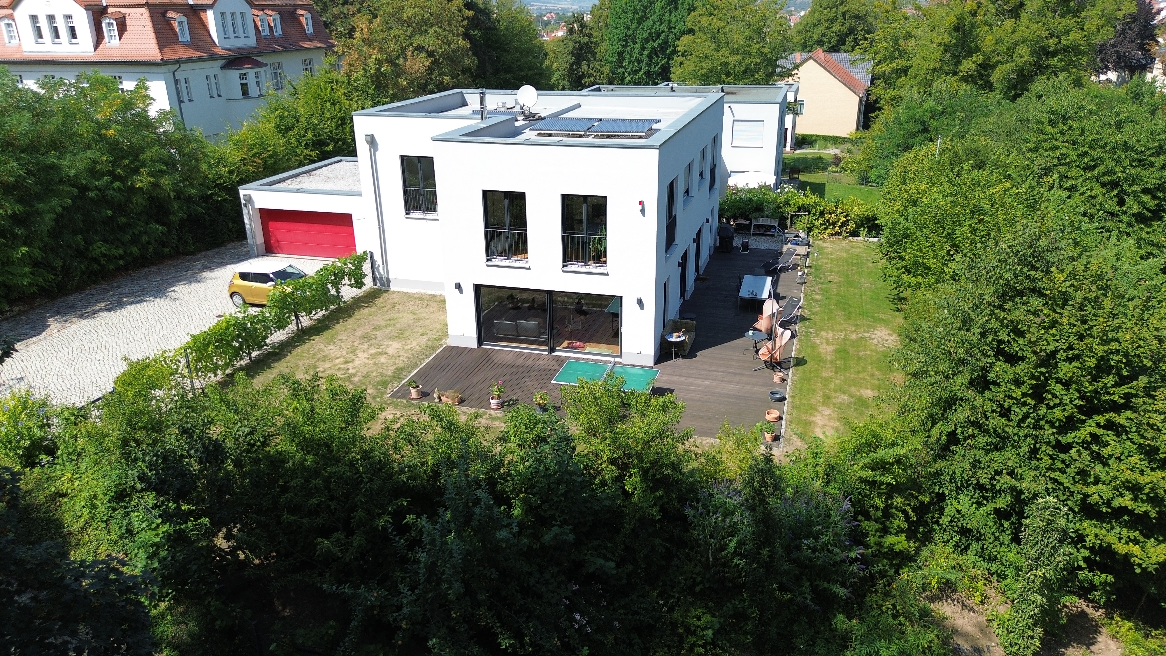 Aerial view of a modern white house with a red garage door, solar panels, and a wooden deck surrounded by lush green trees.