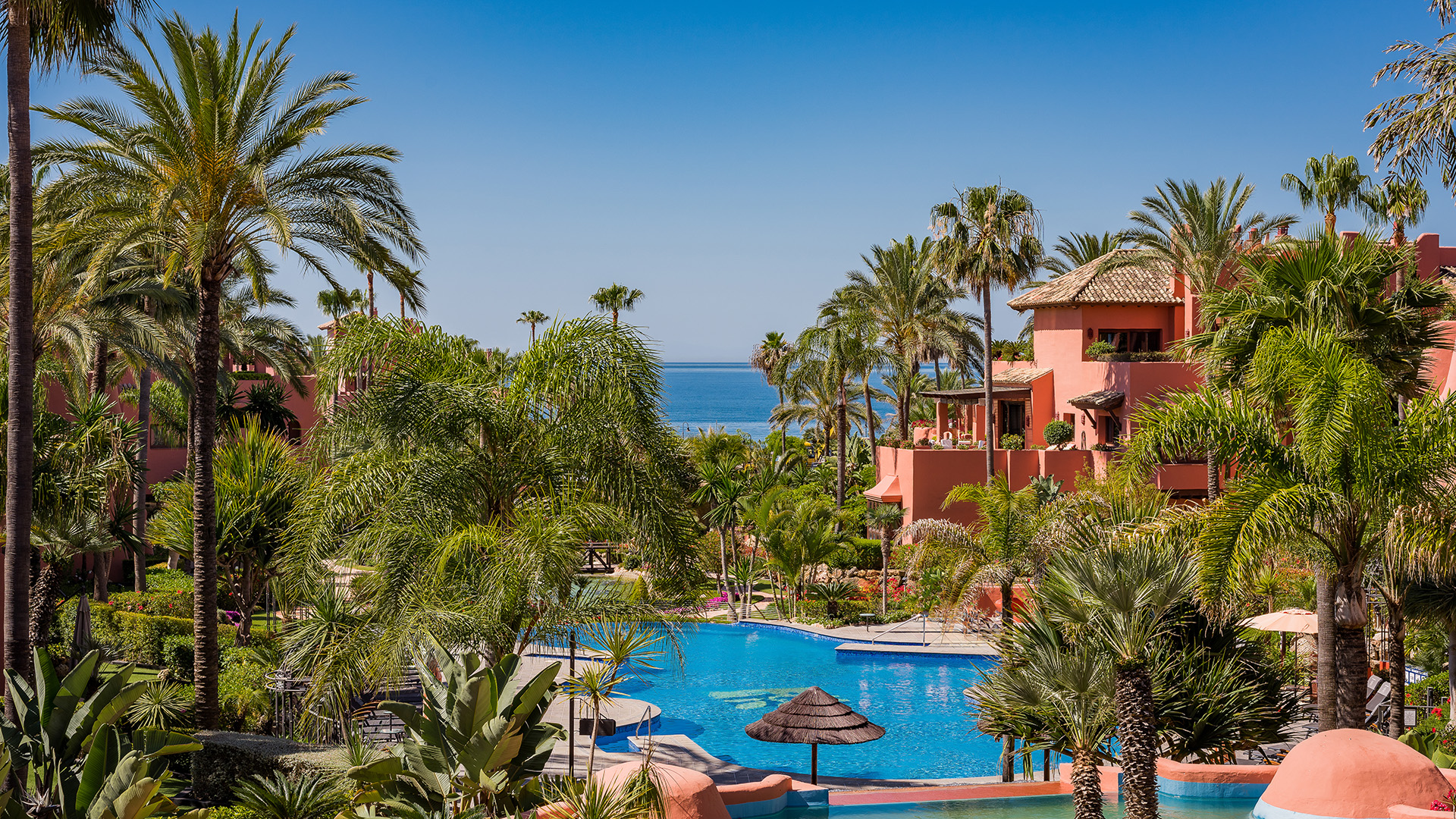 A tropical resort with a blue swimming pool, lush palm trees, and terracotta buildings overlooking the distant ocean.