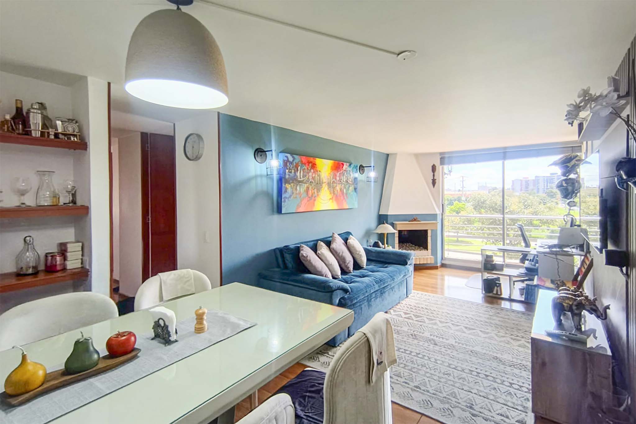 Bright living room with blue accent wall, fireplace, and large windows overlooking greenery. Dining table with fruit centerpiece in foreground.