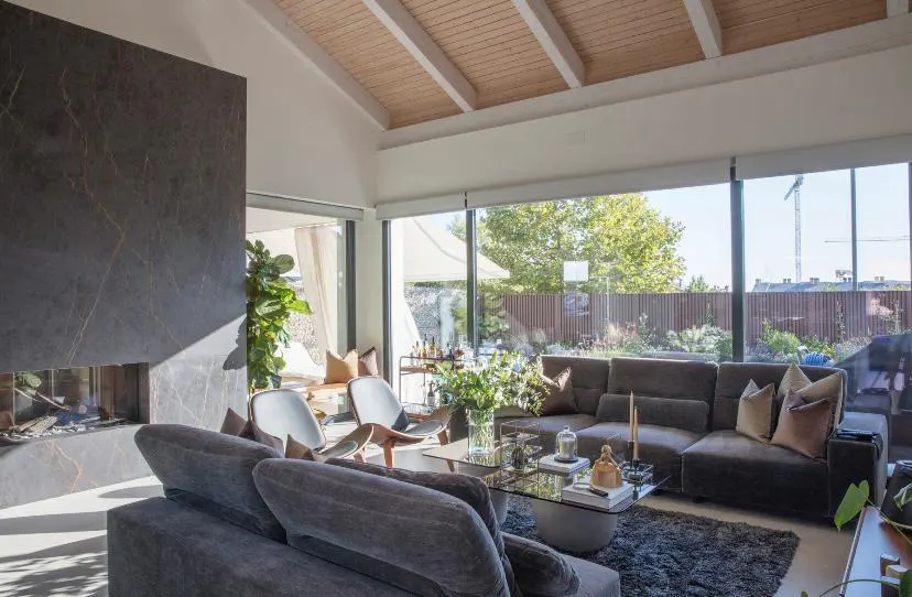 Bright living room with a gray sectional sofa, glass coffee table, and a stone fireplace. Large windows offer a view of the outdoor greenery.