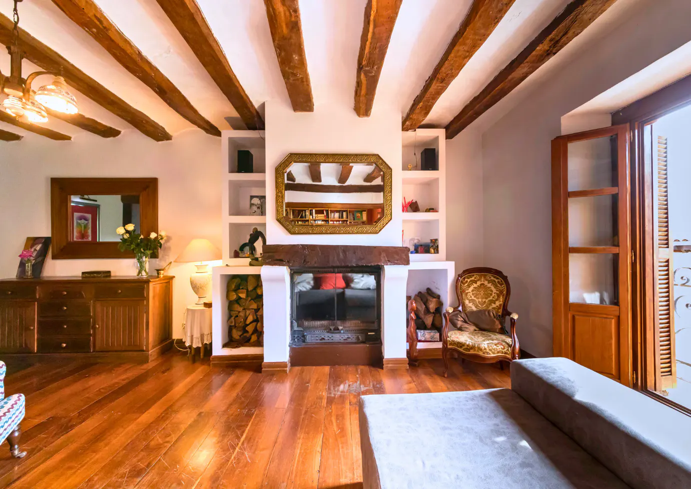 Living room with exposed wood beams, fireplace, and hardwood floors. Built-in shelves flank the fireplace. A brown couch is in the foreground.