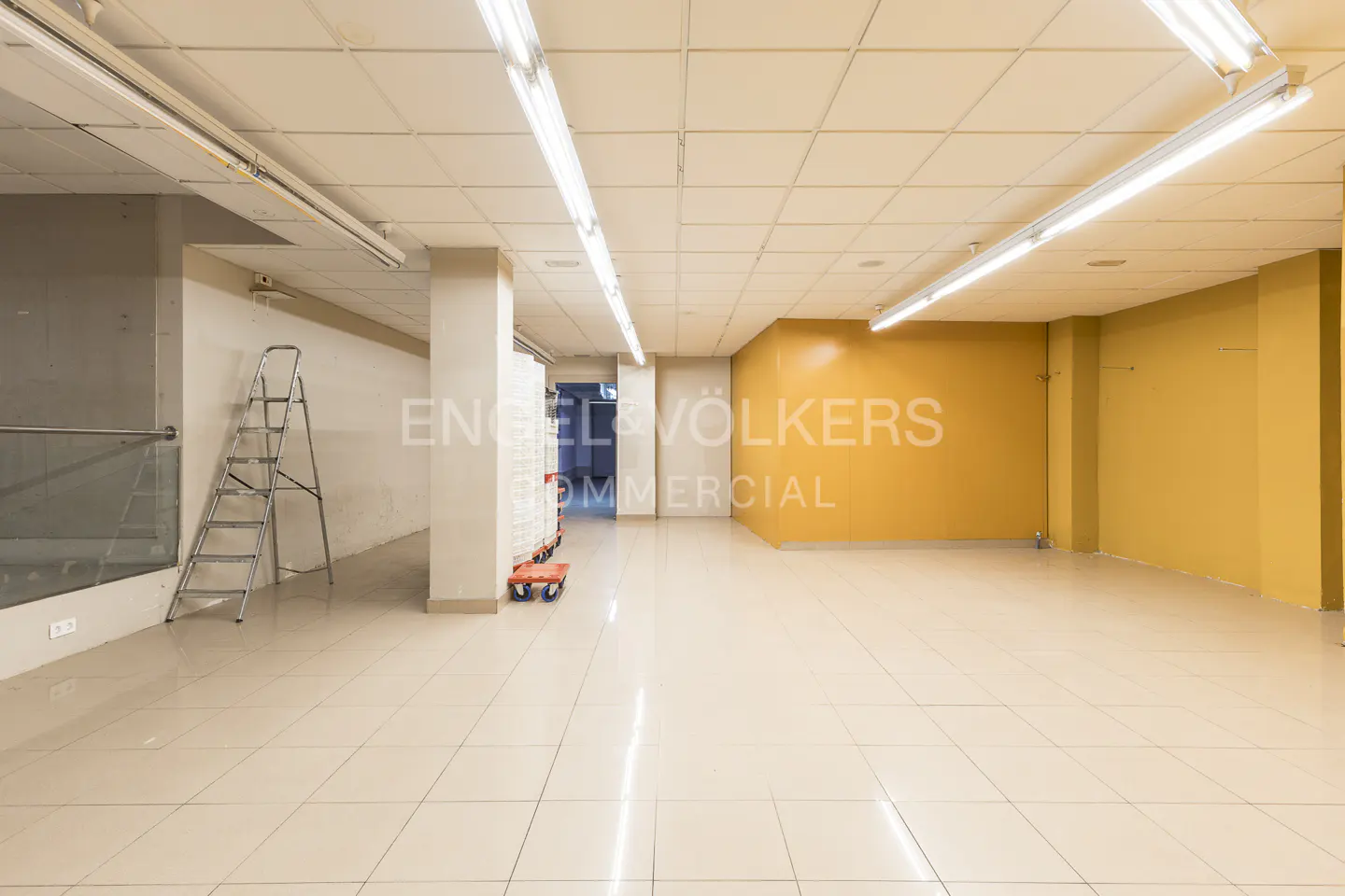 Bright, empty commercial space with beige tile floor, yellow accent wall, and fluorescent lighting. A ladder stands near a glass partition.