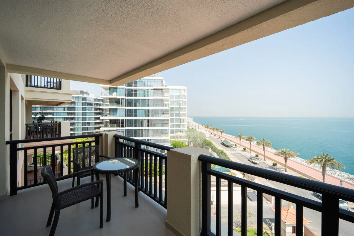 Covered balcony with a small table and two chairs, overlooking a coastal road, palm trees, the ocean, and modern buildings.