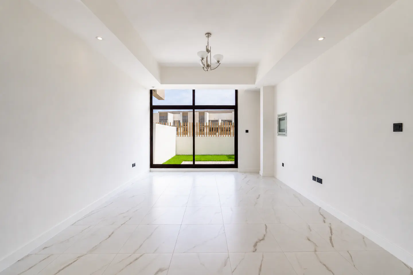 Bright, empty room with white walls, marble floors, and a black-framed window showing a green lawn and white fence. A chandelier hangs from the ceiling.