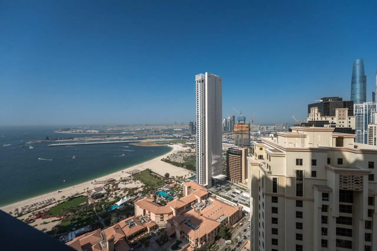 Aerial view of Dubai's skyline, featuring a beach, ocean, and modern buildings under a clear blue sky.