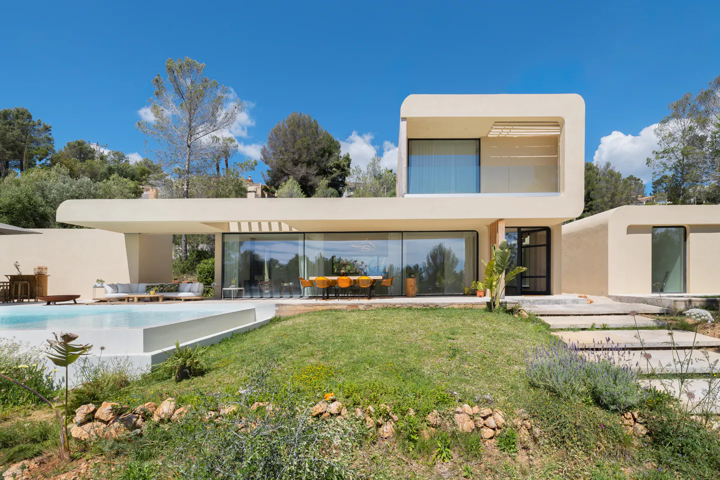 Modern beige house with large windows, a pool, and green lawn under a blue sky. Trees surround the property.