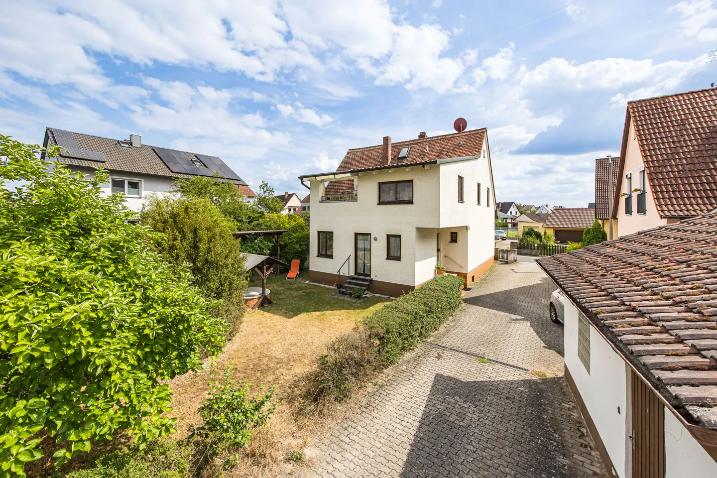 Two-story white house with a brown roof, small yard, and paved driveway on a sunny day.