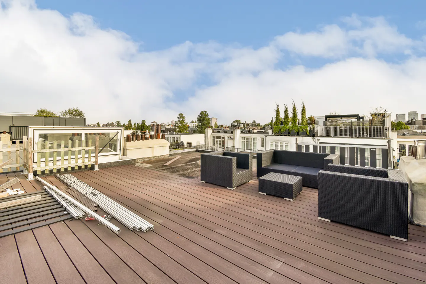 Rooftop patio with brown wood flooring, black wicker furniture, and a view of surrounding buildings under a cloudy sky.