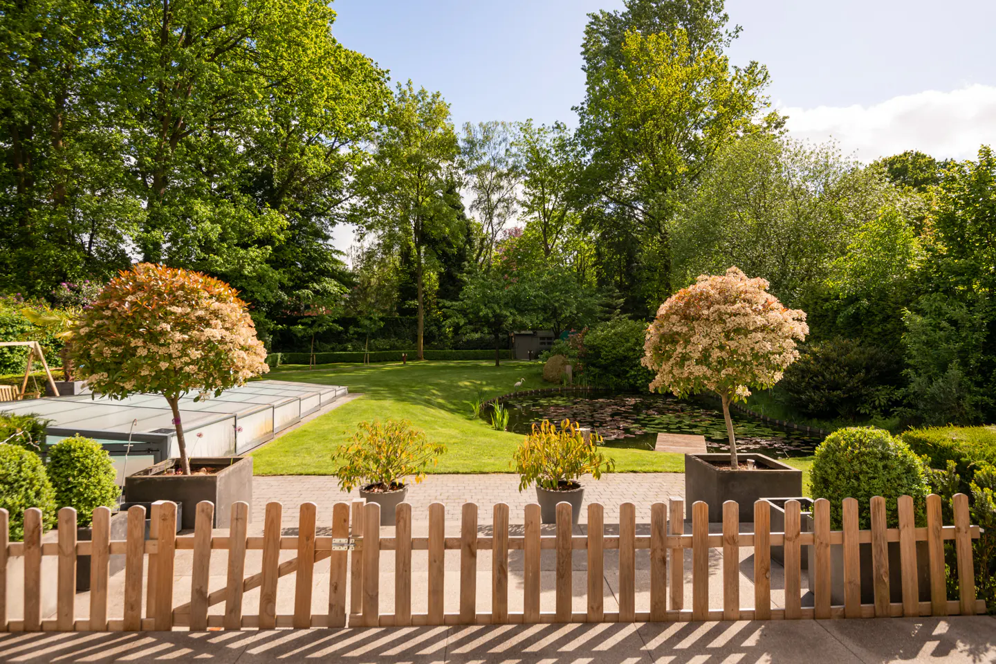 View of a lush green garden with trees, lawn, pond, and potted plants behind a light brown wooden fence.