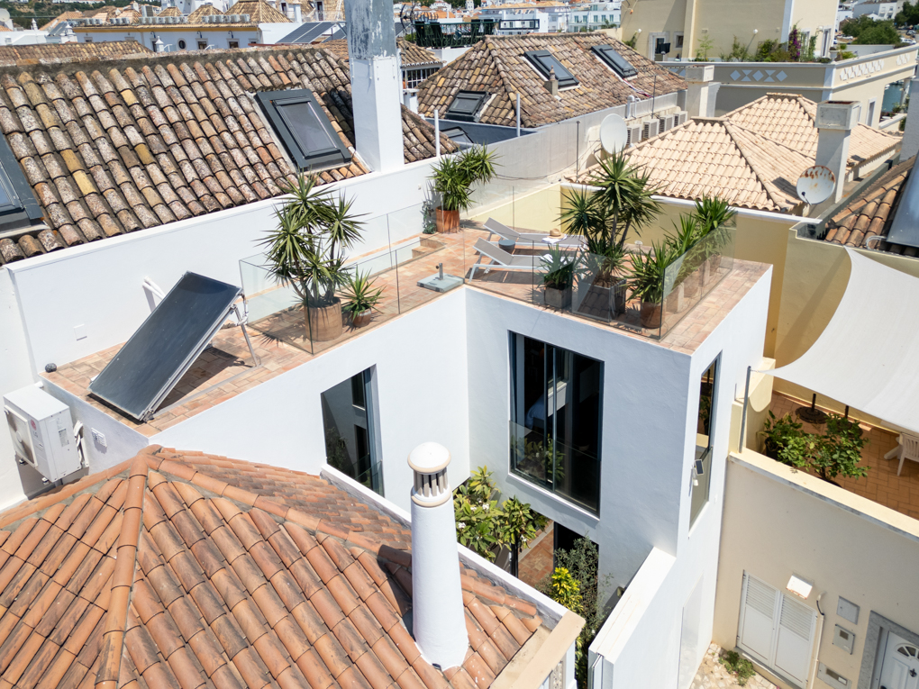 High-angle view of a modern white house with a rooftop terrace, potted plants, lounge chairs, and a solar panel.