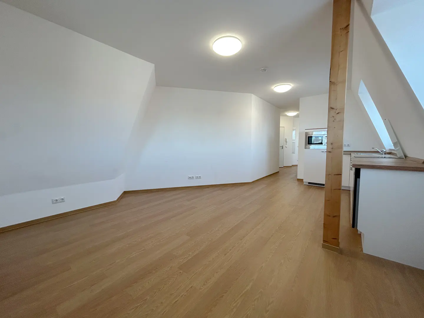 Bright, empty apartment with wood floors, white walls, and a partial kitchen view. A wooden beam supports the sloped ceiling.