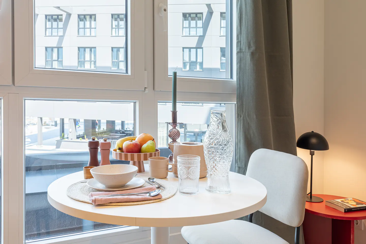 Bright apartment interior with a round white table set for a meal, a white chair, and a window view of a modern building.