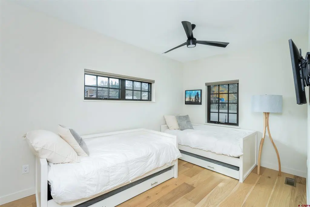 A bright bedroom with two white twin beds, white walls, and light wood floors. A black ceiling fan and two windows add contrast.
