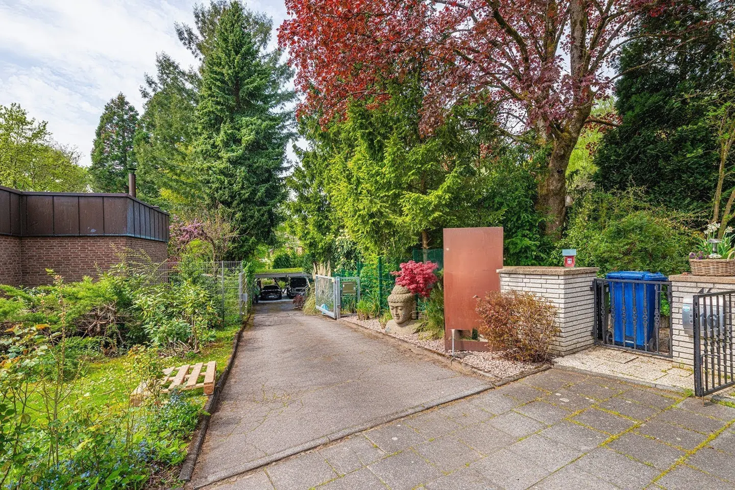 View of a driveway leading to a house, surrounded by lush greenery and trees. A Buddha statue and blue trash can are visible.
