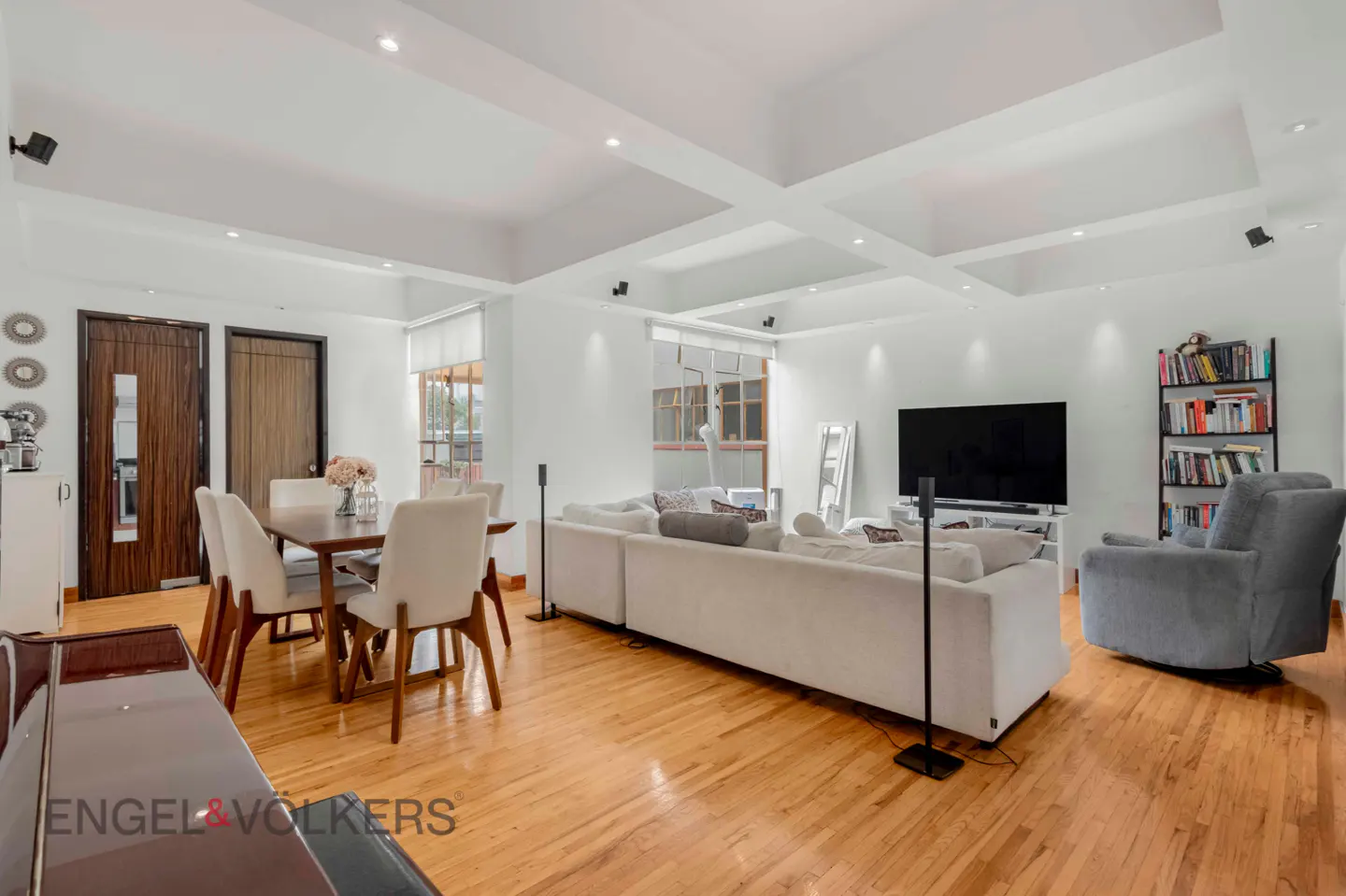 Bright living room with hardwood floors, white walls, and coffered ceiling. Features a dining table, white sofa, TV, and bookshelf.