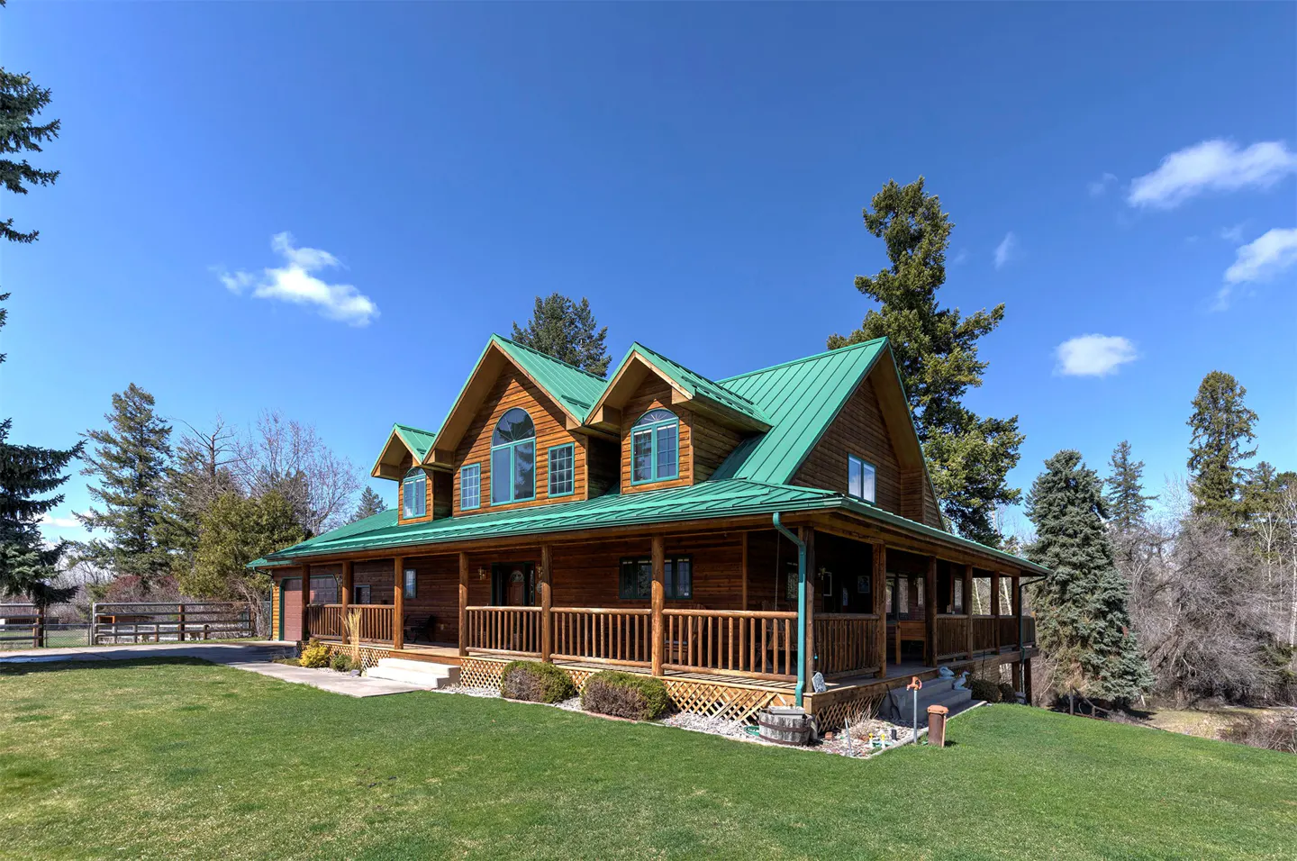 A two-story log home with a green metal roof and a wrap-around porch on a green lawn under a blue sky.