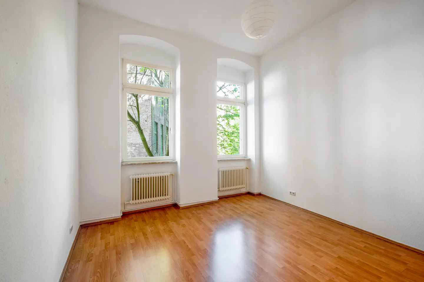 Empty room with wood floor, white walls, and two windows with radiators. A white paper lantern hangs from the ceiling.