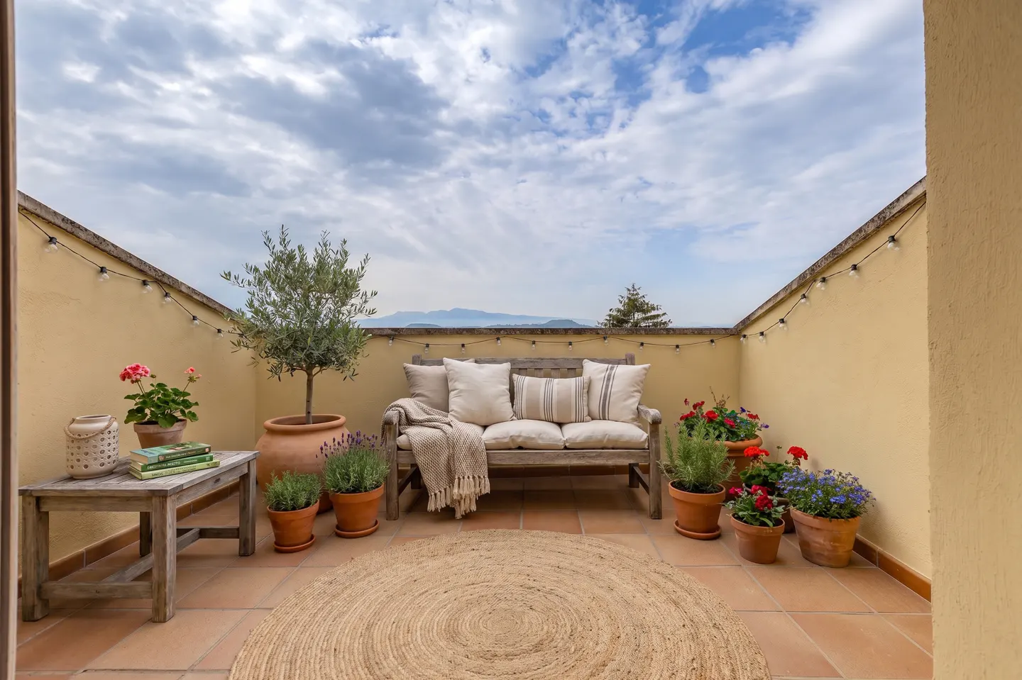 Outdoor patio with a wooden bench, pillows, plants in terracotta pots, and a round jute rug. String lights hang on the yellow walls.