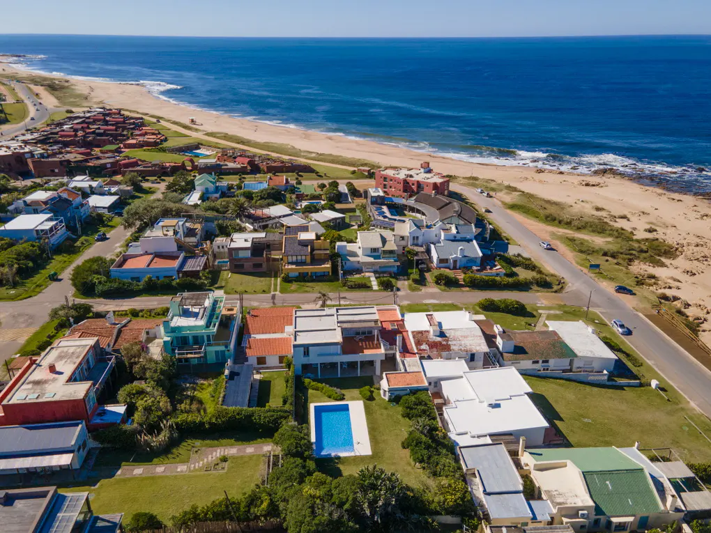 Aerial view of a coastal neighborhood with houses, a swimming pool, and a sandy beach along the blue ocean.