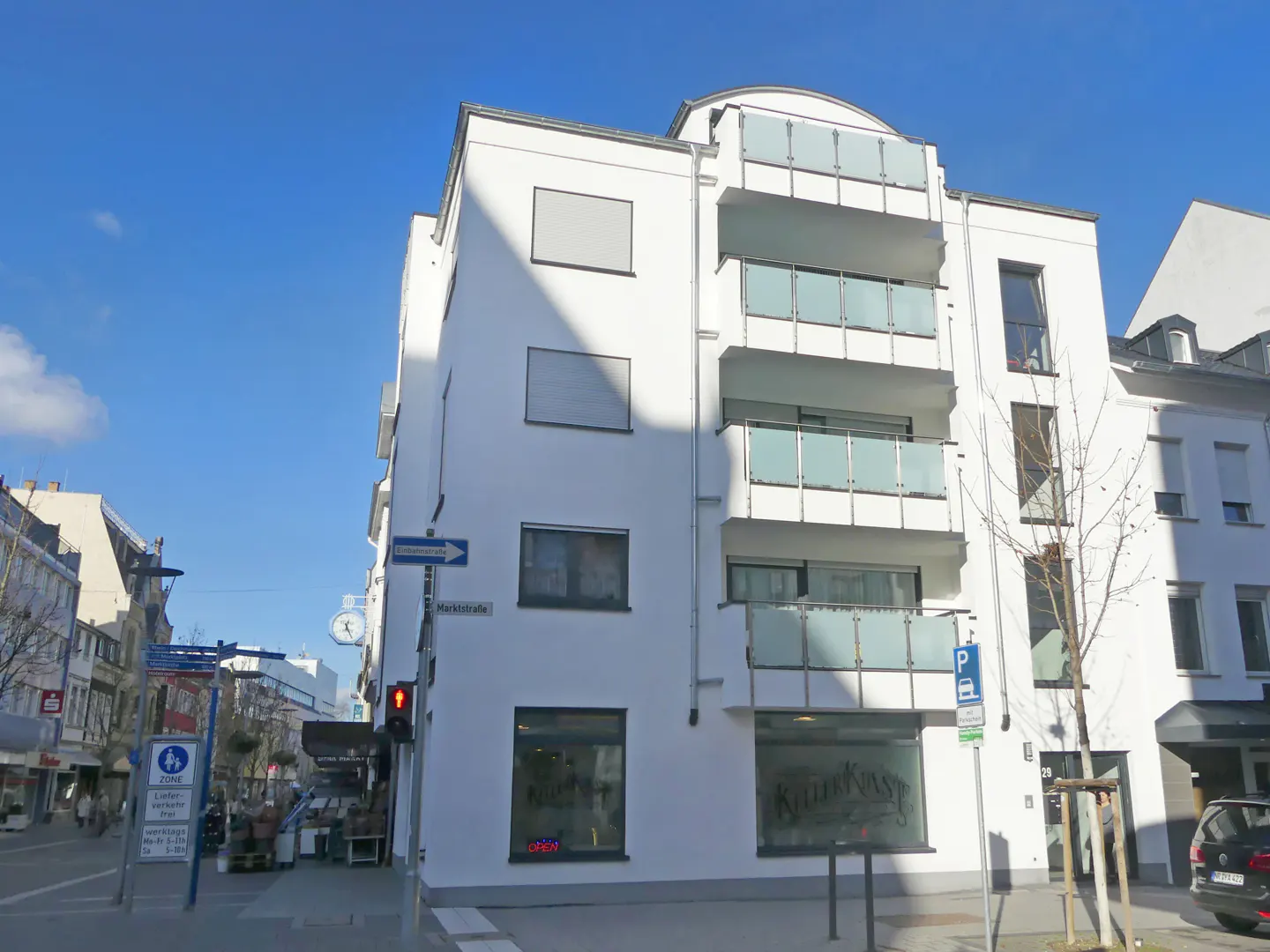 Exterior view of a modern white building with glass balconies on a sunny day. Street signs and shops are visible.