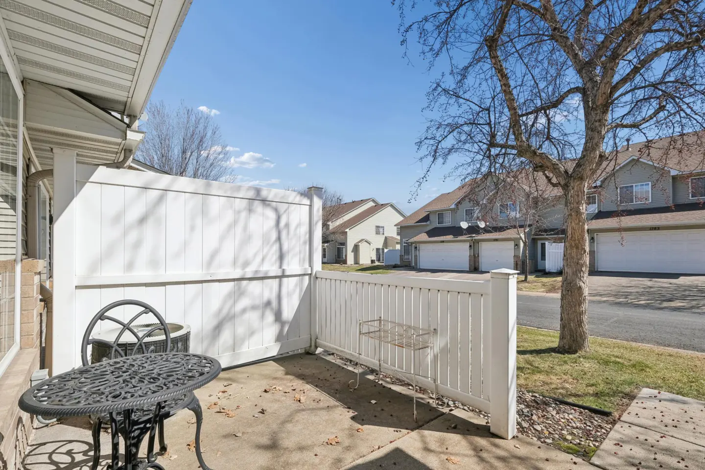 Outdoor patio with black metal table and chairs, white fence, and townhouses in the background on a sunny day.