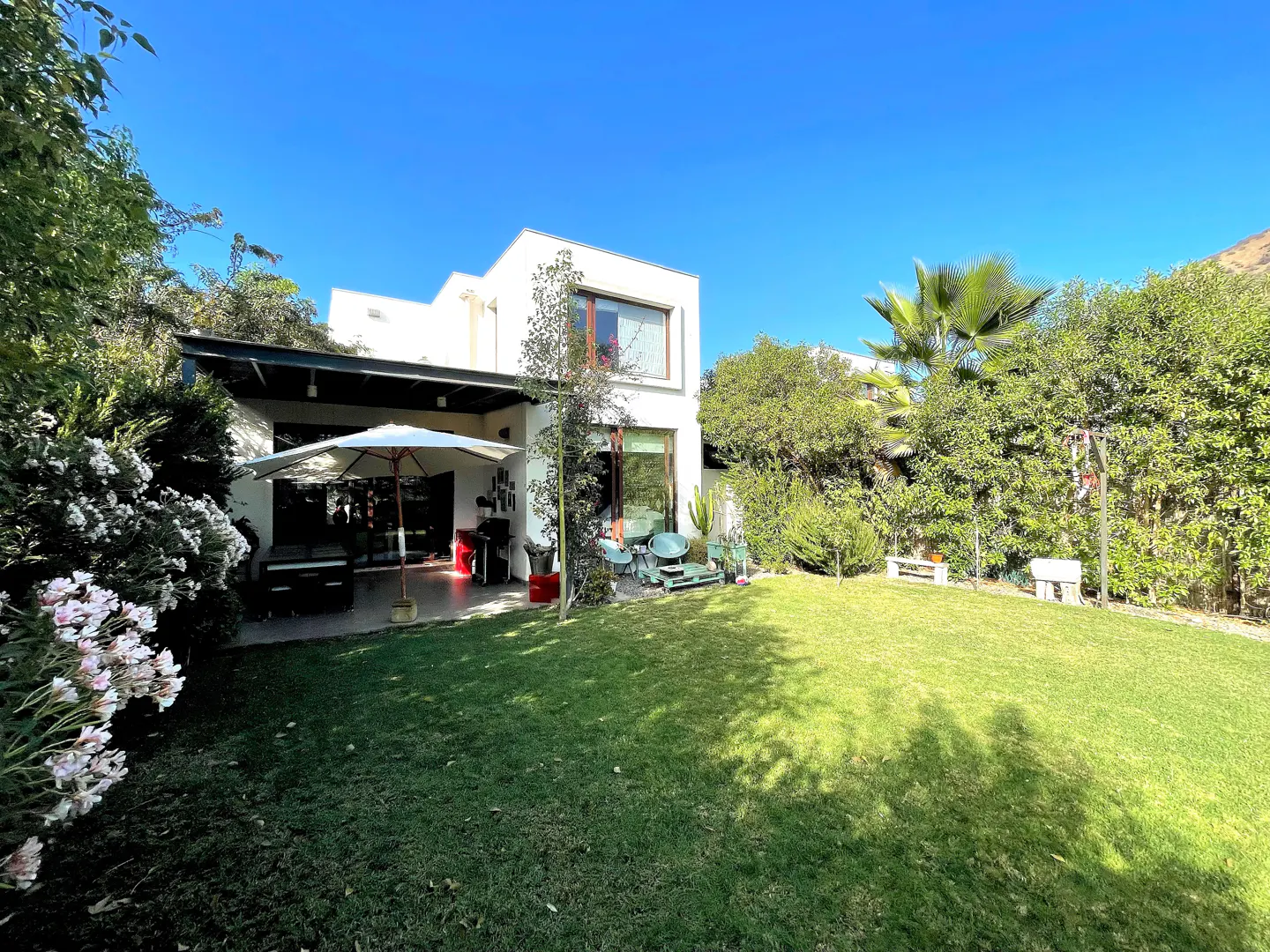 Modern white house with a patio, white umbrella, and lush green lawn under a clear blue sky.