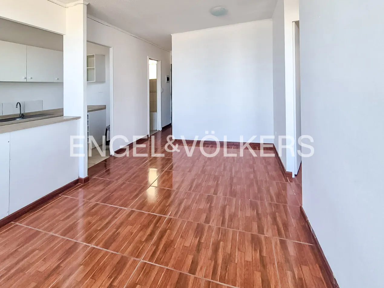 An interior shot of a bright, empty apartment with brown tile floors and white walls. A kitchen area is visible on the left.