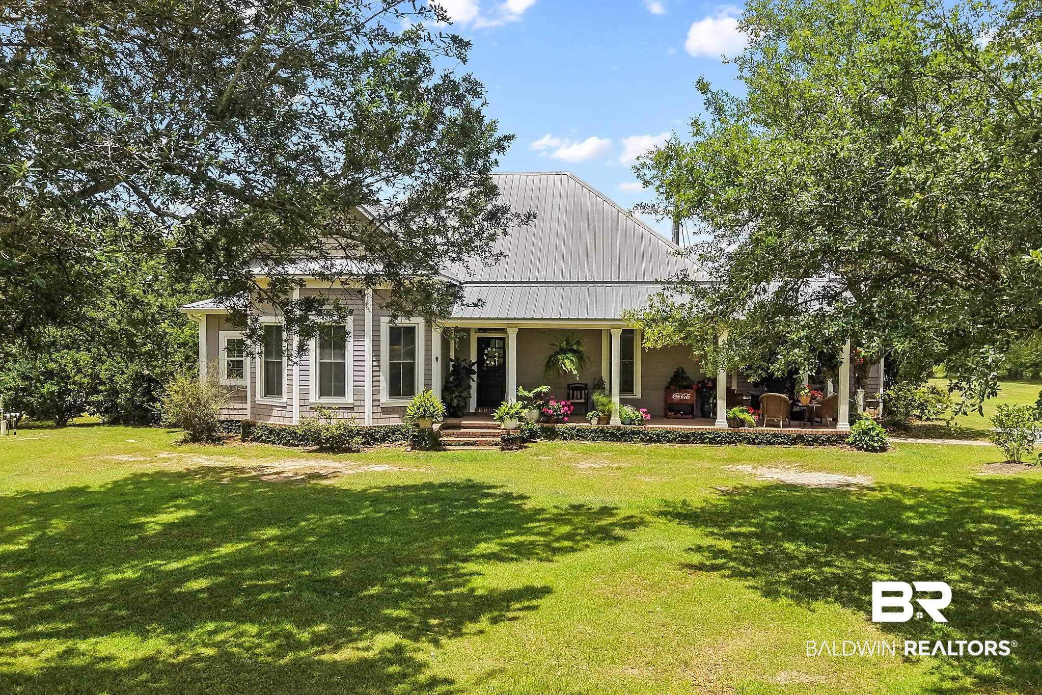 Single-story gray home with a metal roof, large front porch, and lush green lawn under mature oak trees.
