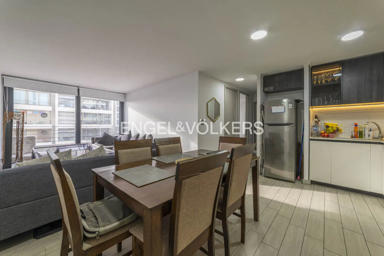 A modern apartment interior with a dining table, gray sofa, and stainless steel kitchen appliances.