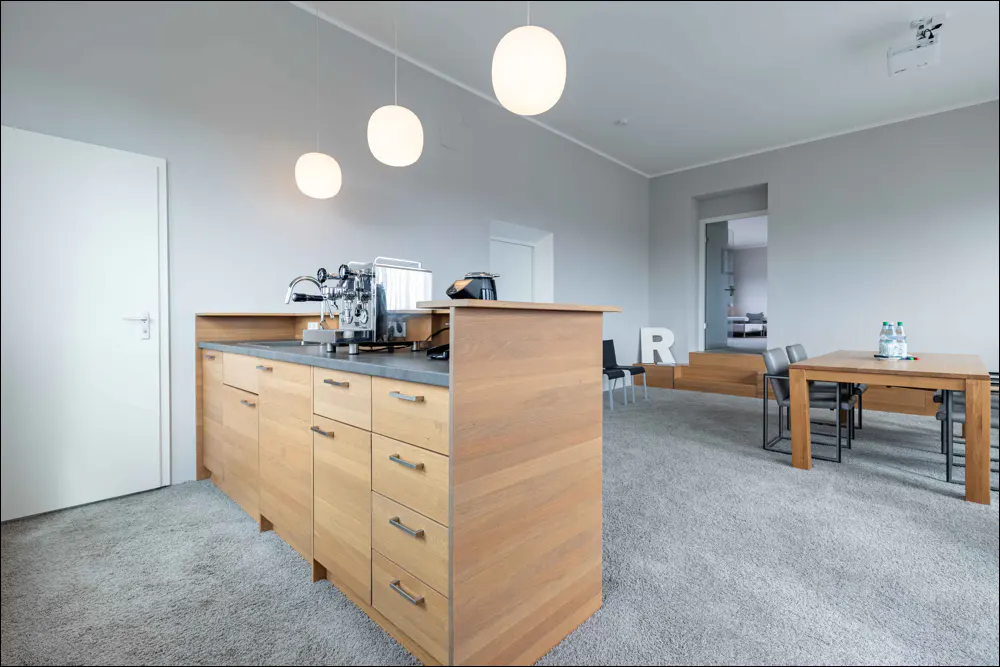 Bright, modern office space with a wooden kitchen island, stainless steel espresso machine, and a wooden table with black chairs.