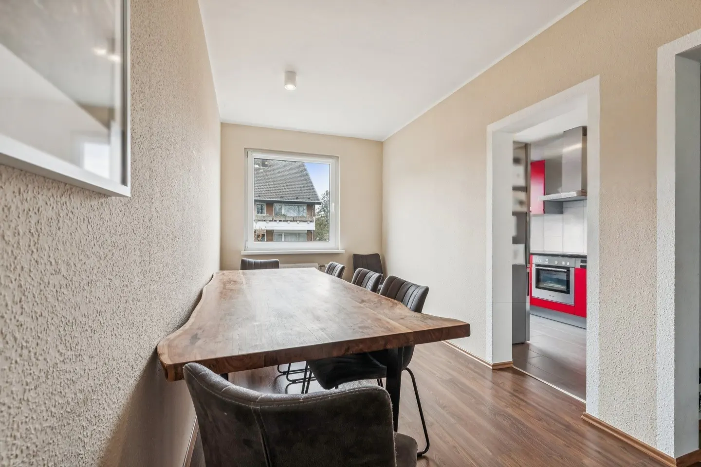 Dining room with a long wooden table, gray chairs, and a view into a red and gray kitchen.