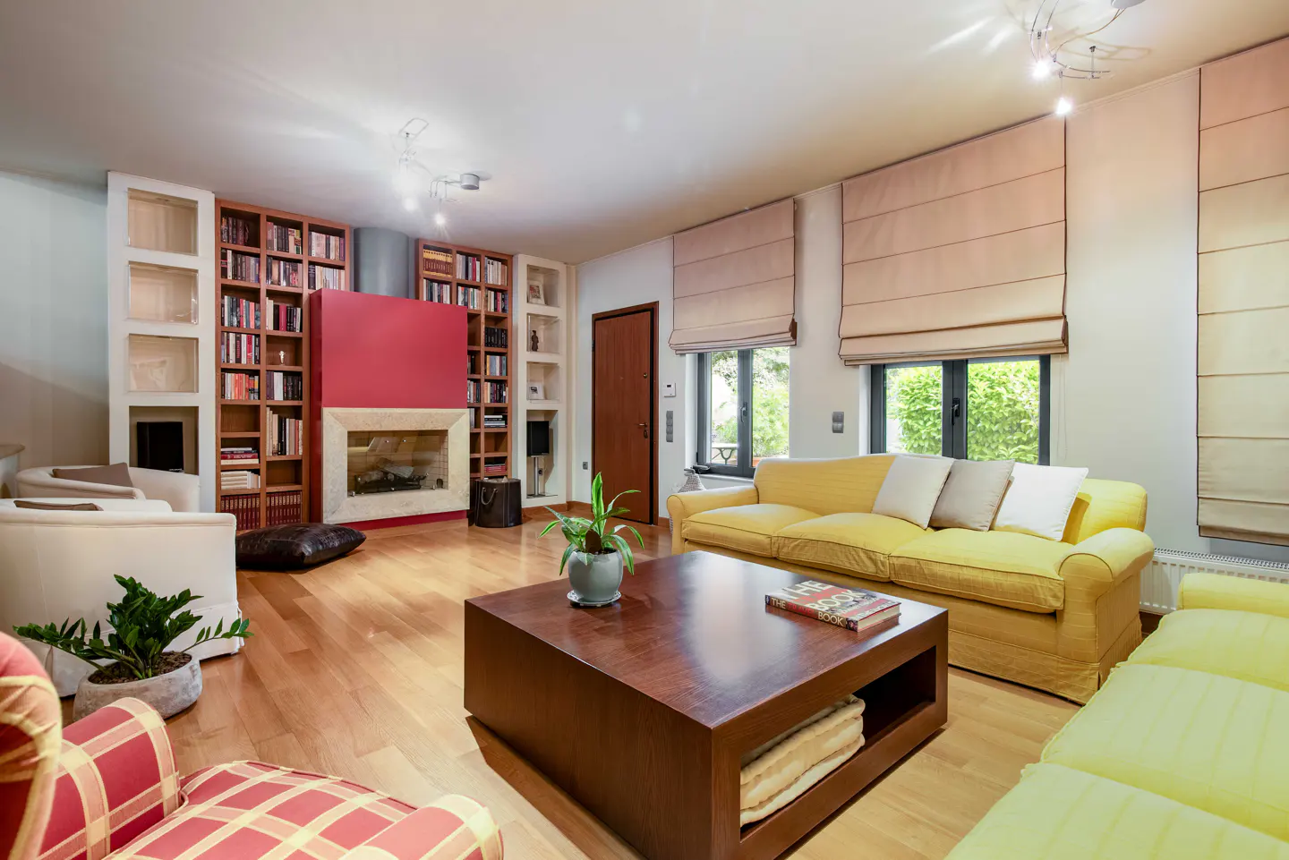 Bright living room with hardwood floors, yellow sofa, and a dark wood coffee table. A fireplace is flanked by bookshelves. Windows have tan roman shades.