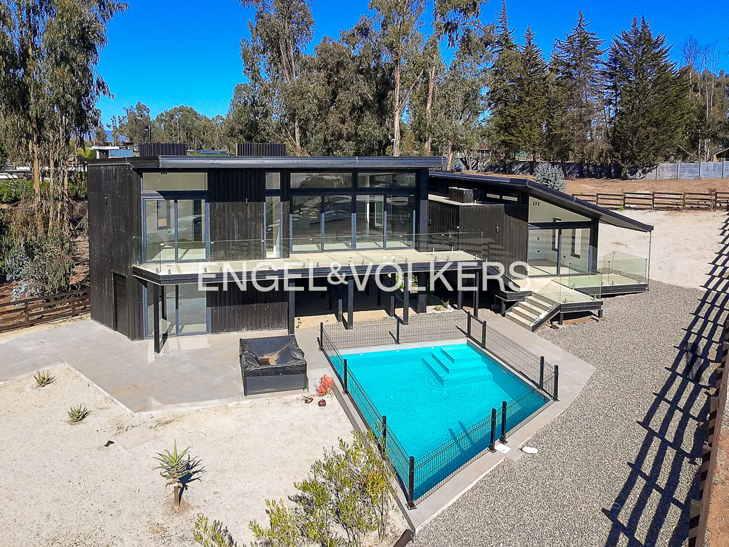 Modern black house with a blue pool, surrounded by trees and gravel. Engel & Volkers logo is visible on the house.