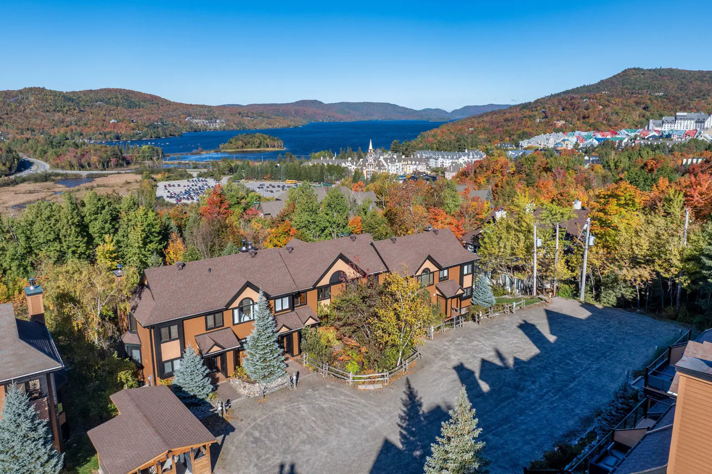 Aerial view of a brown townhouse complex with a lake and mountains in the background. Fall foliage colors the trees.