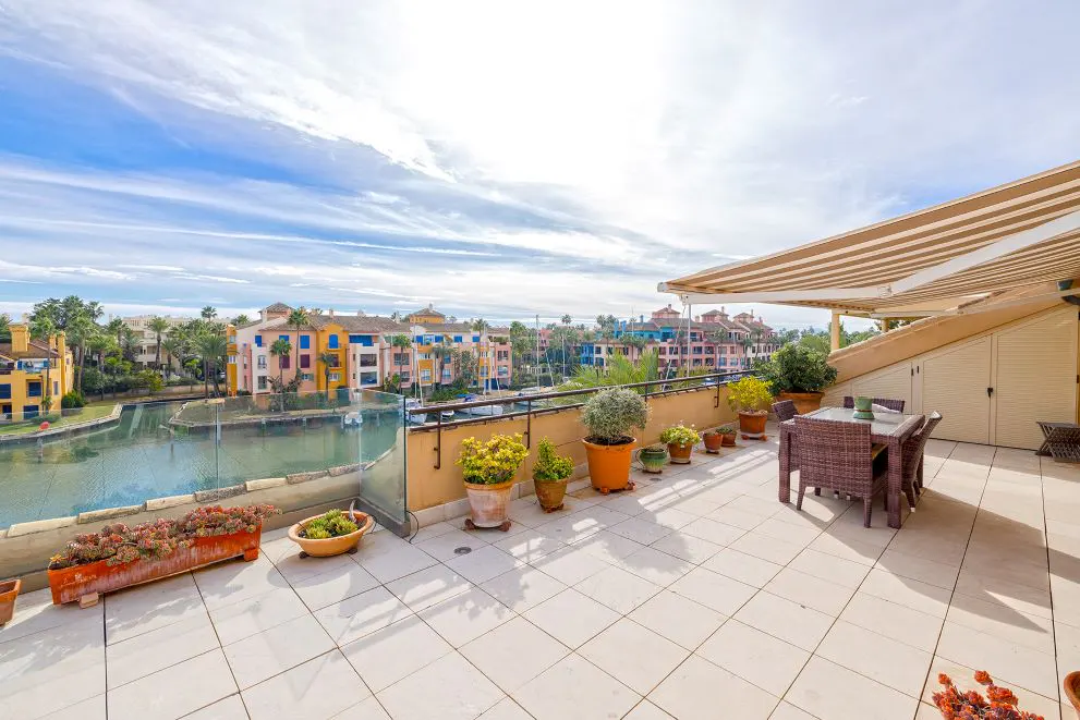 A sunny patio with potted plants, a table, and a view of colorful buildings and a canal.