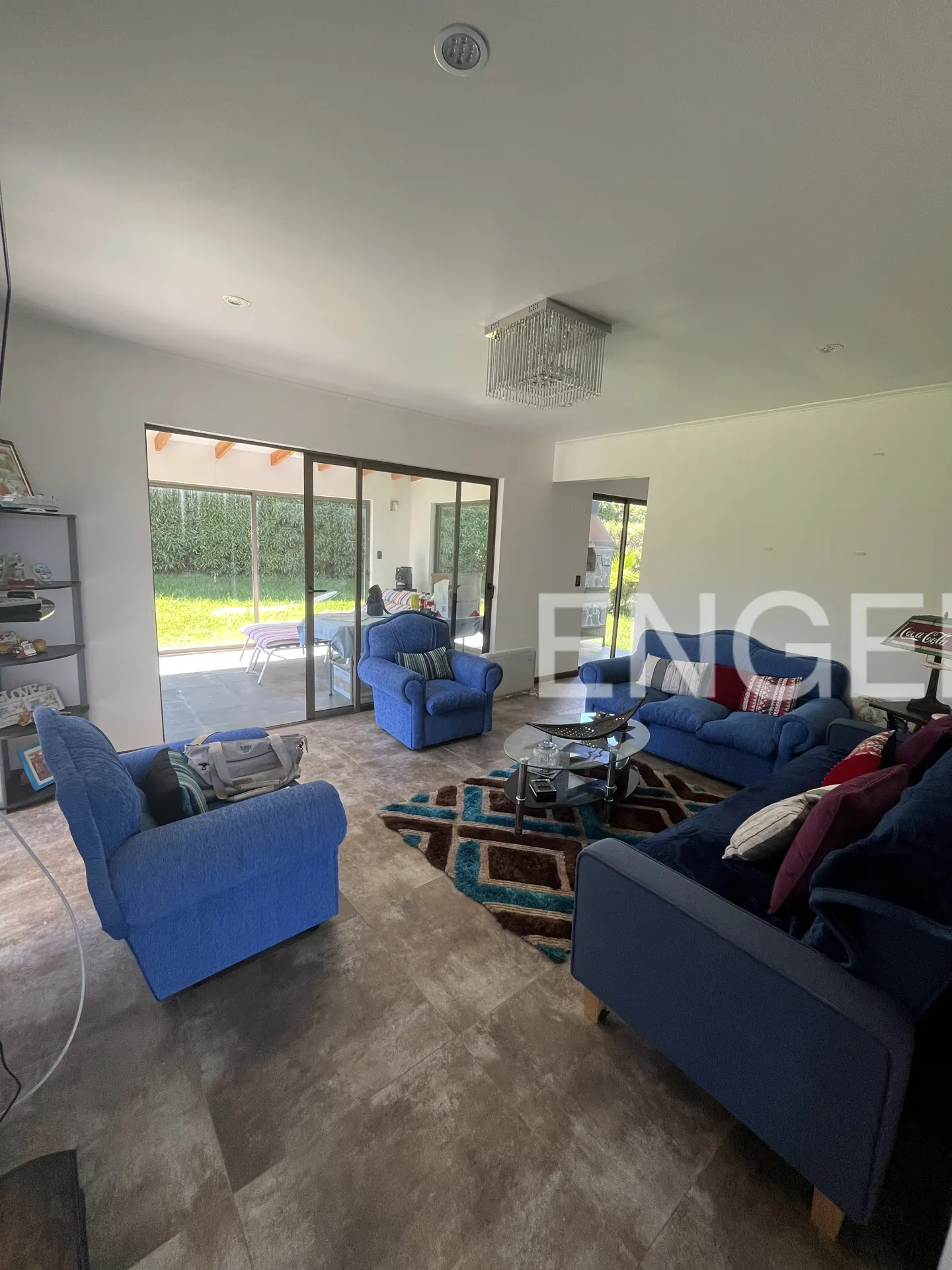 Bright living room with blue sofas, armchairs, and a patterned rug. Sliding glass doors lead to a patio and green lawn. Chandelier hangs from the white ceiling.