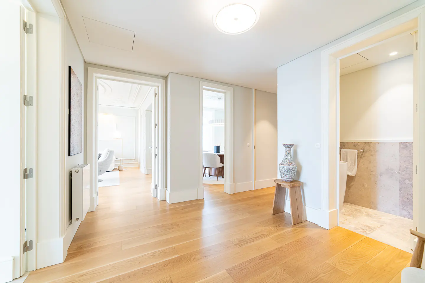 Hallway with light wood floors, white walls, and open doorways. A decorative vase sits on a small wooden stool.