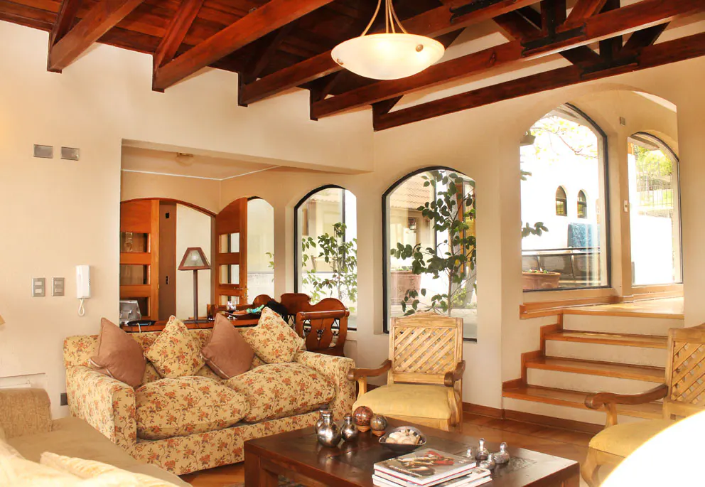 Living room with floral sofa, wood beams, and arched windows. A wooden coffee table sits in front of the sofa. Stairs lead to another room.