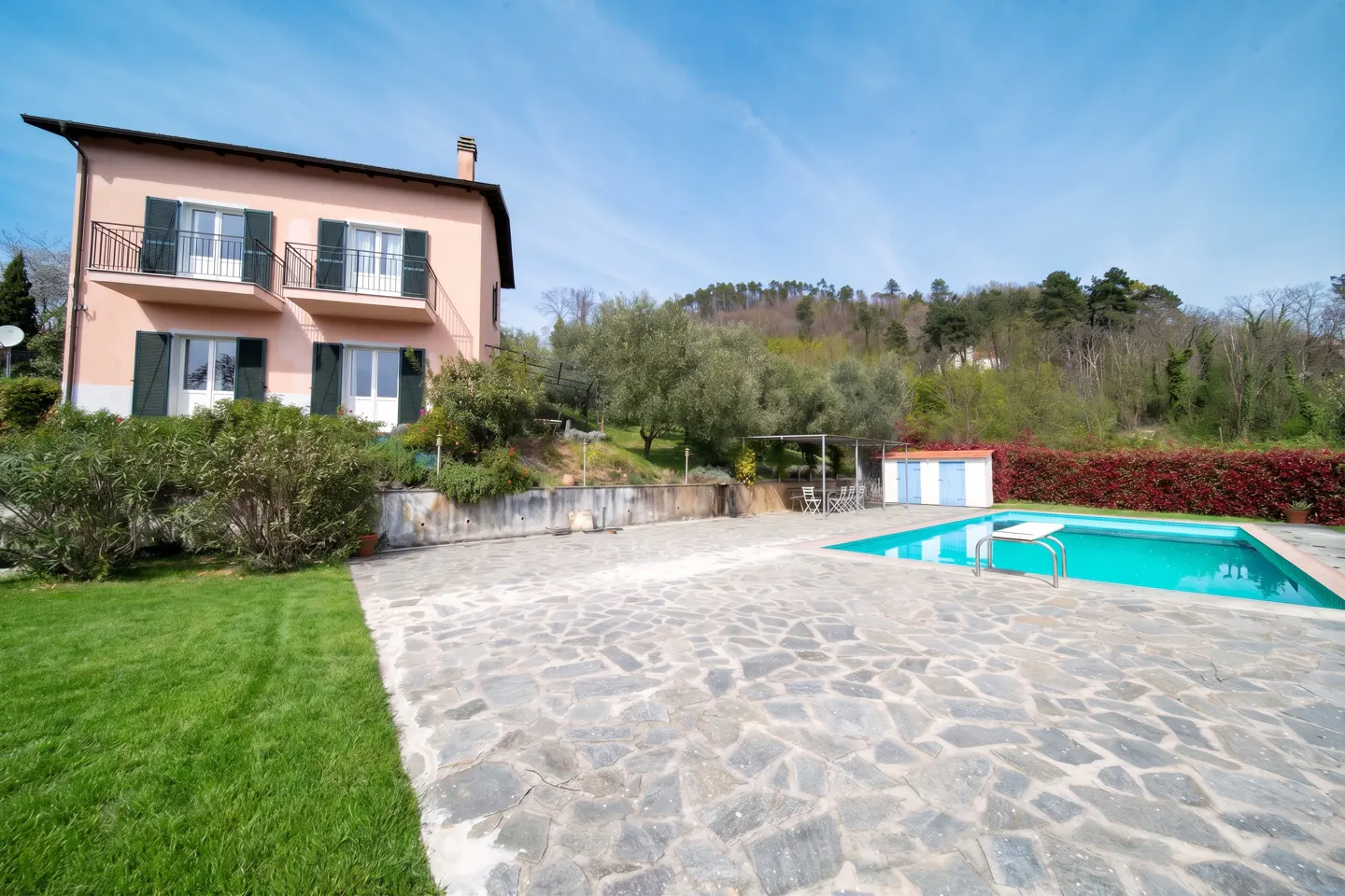 Two-story pink house with green shutters, a pool, and a stone patio on a sunny day.