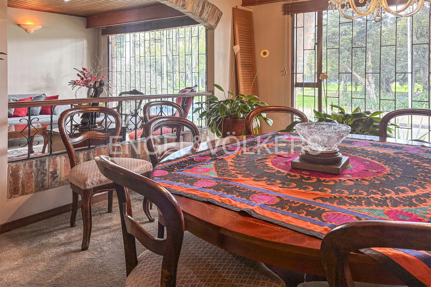 A dining room with a wooden table covered in a colorful patterned cloth, surrounded by wooden chairs. A crystal bowl sits in the center.