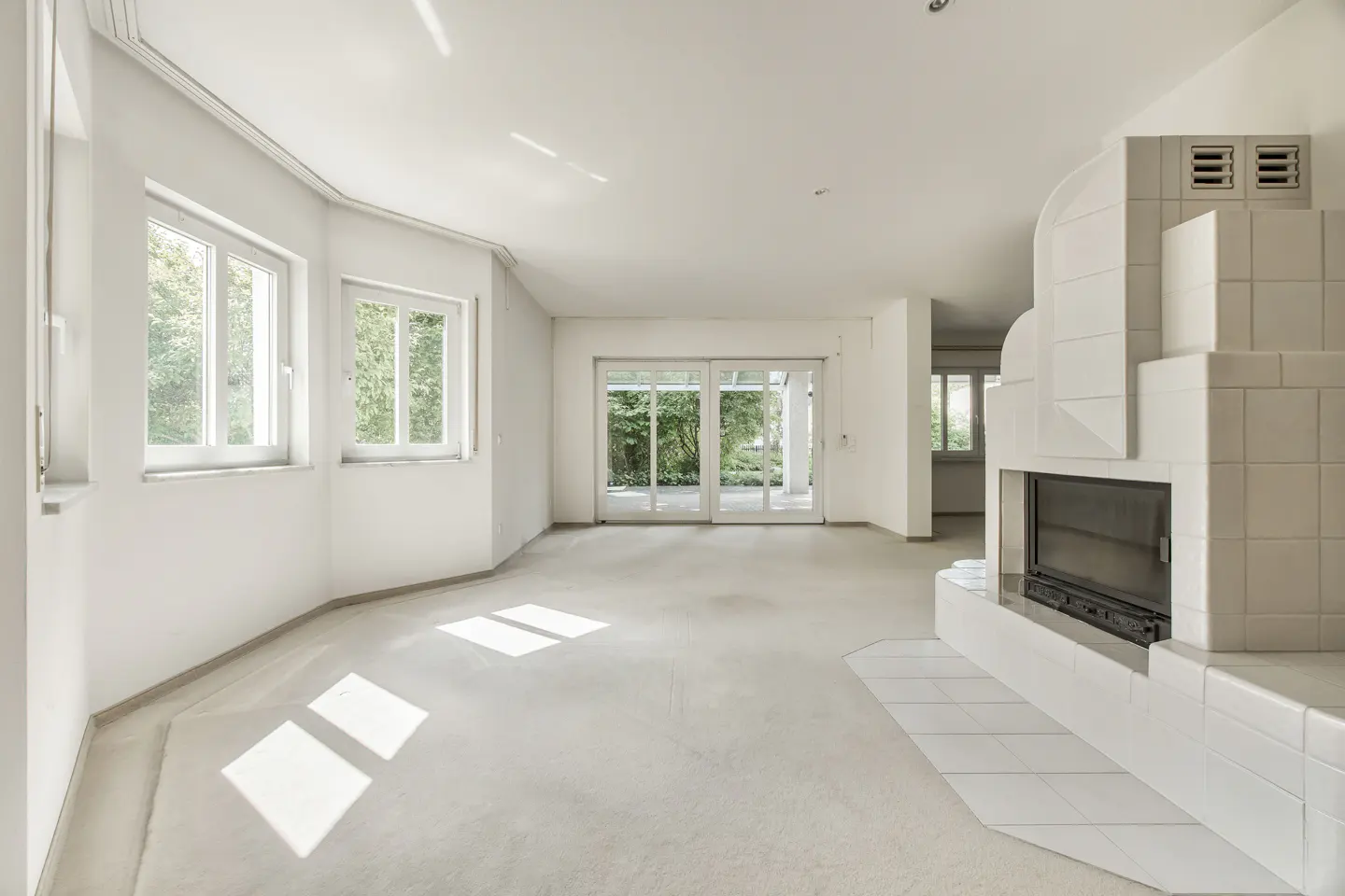 Bright, empty living room with white walls, light flooring, large windows, sliding glass doors, and a white tiled fireplace.
