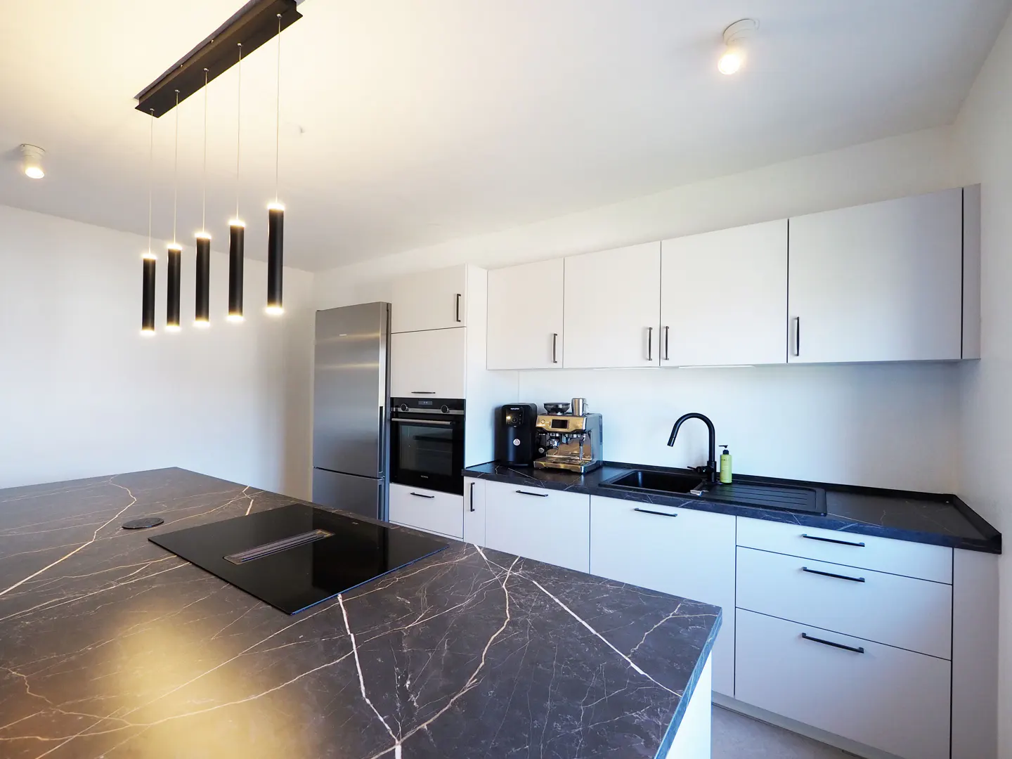 Bright kitchen with white cabinets, black countertops, and a marble island with a built-in cooktop. Stainless steel appliances and modern pendant lights.