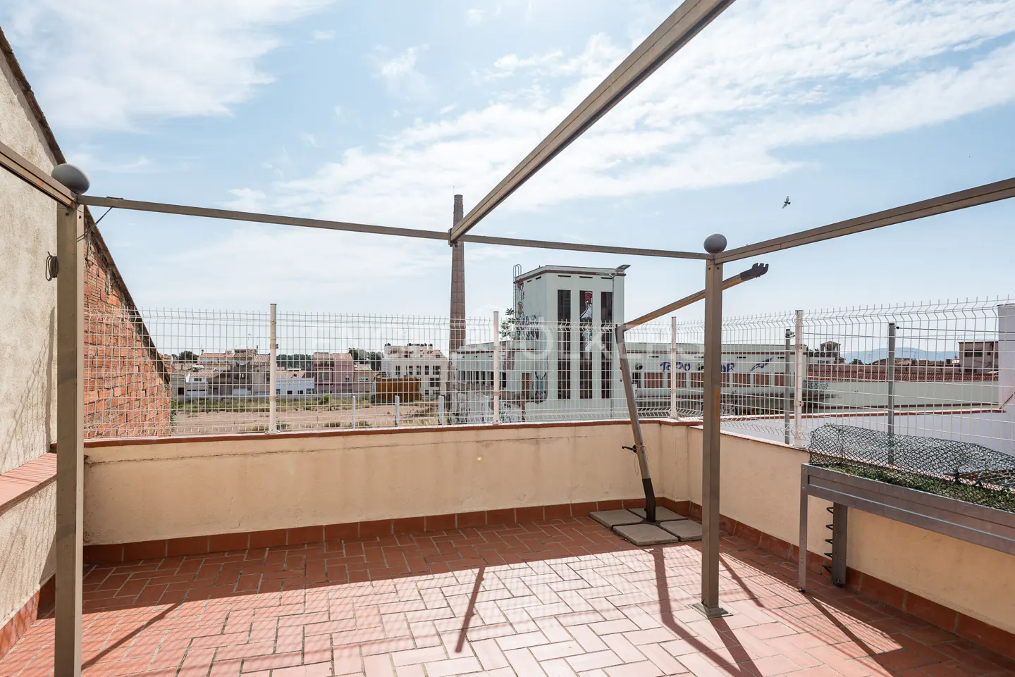 A rooftop terrace with red brick tiles, a metal pergola, and a white wire fence overlooking a town with a tall brick chimney.
