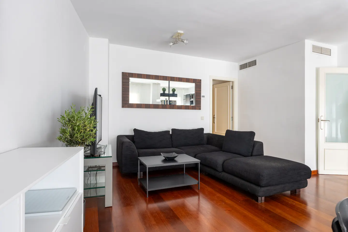 Living room with dark gray sectional sofa, black coffee table, and wood floors. A mirror hangs above the sofa.