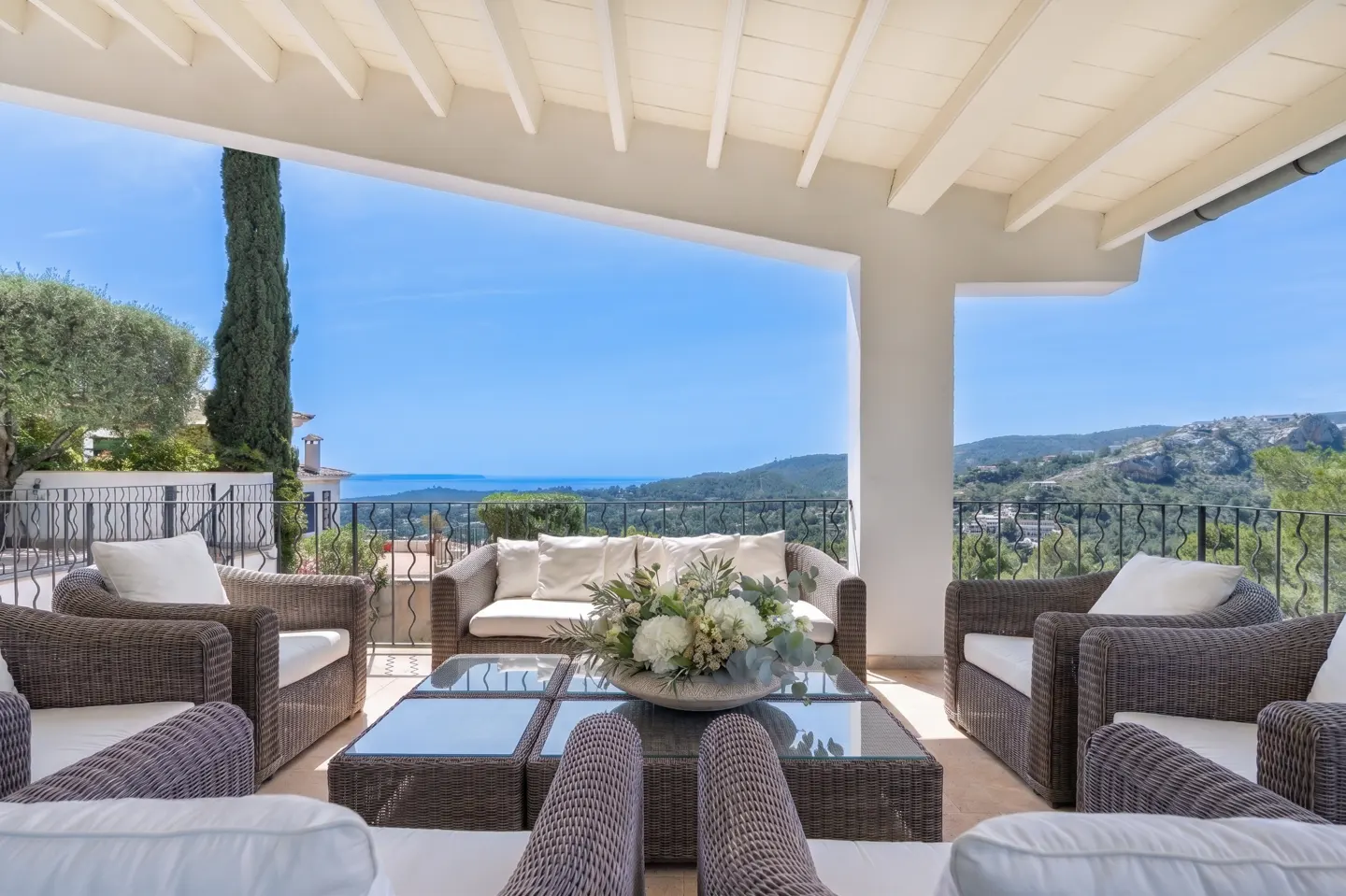 Outdoor patio with wicker furniture, white cushions, and a glass-topped coffee table. Ocean and green hills visible in the background.