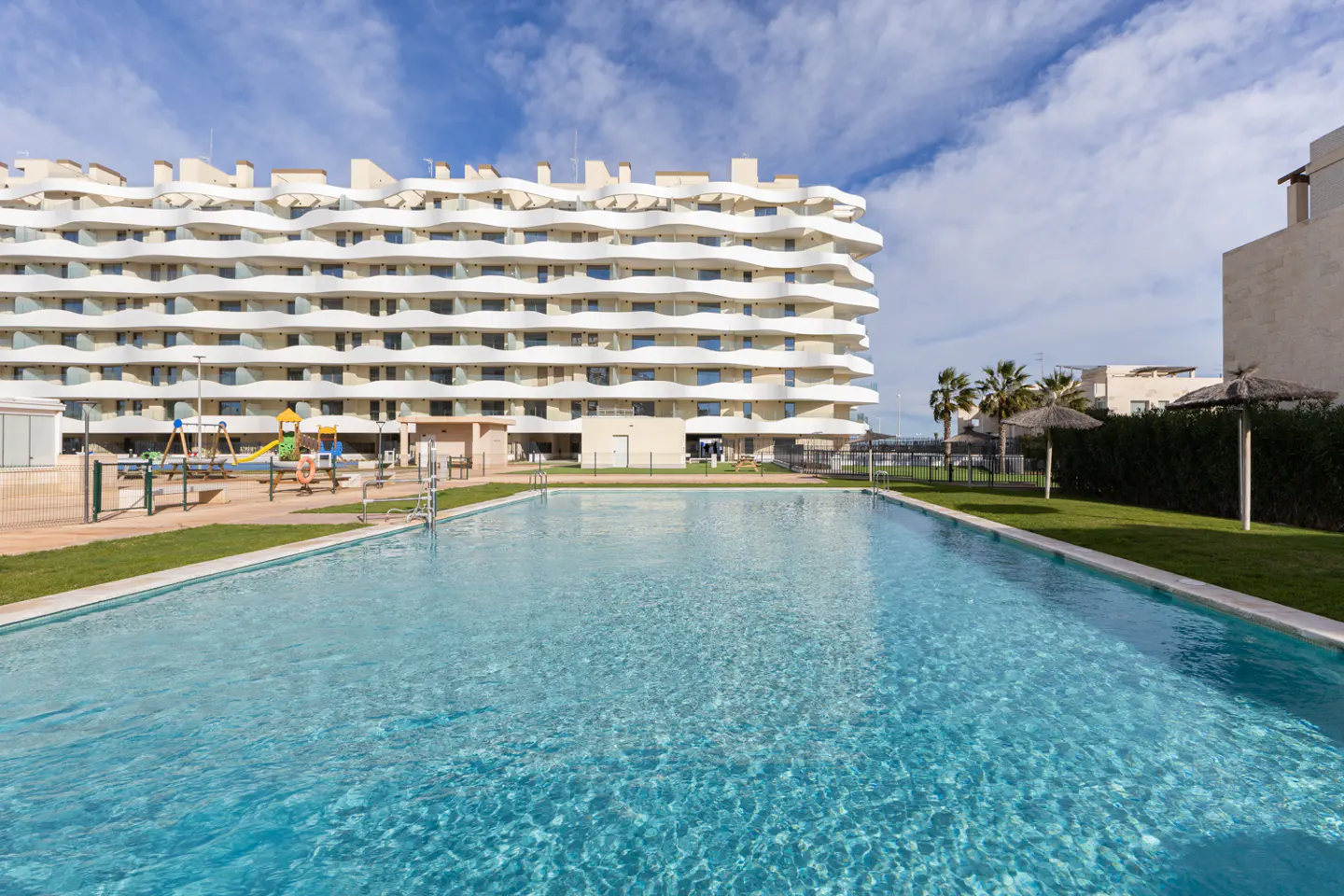 A large, turquoise swimming pool in front of a modern, white apartment building with a playground.
